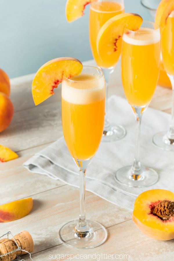 champagne flute filled with peach bellini cocktail, with white foam on the top and a peach slice garnish. Fresh peaches are scattered around the glass and three more peach bellinis are in the background