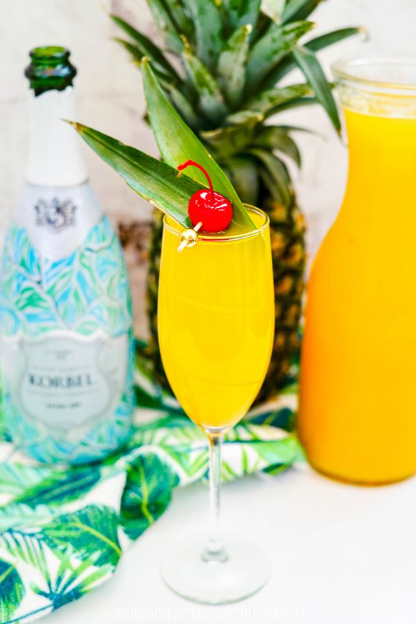 picture of a champagne flute filled with a pineapple mimosa, topped with a pineapple leaf and cherry garnish, with the ingredients needed to make the cocktail in the background