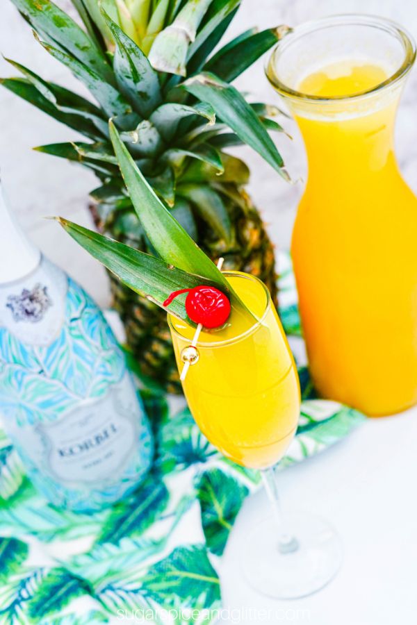 overhead picture of a champagne flute filled with a pineapple mimosa, topped with a pineapple leaf and cherry garnish, with the ingredients needed to make the cocktail in the background