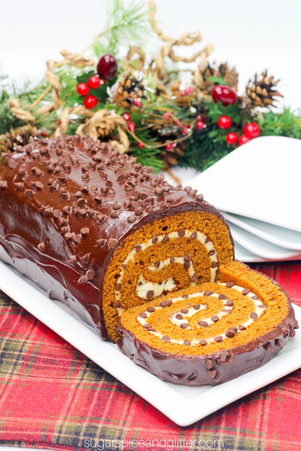 overhead image of a pumpkin cake roll with a swirl of cream cheese frosting inside and a glossy chocolate ganache topping with holiday decorations in the background