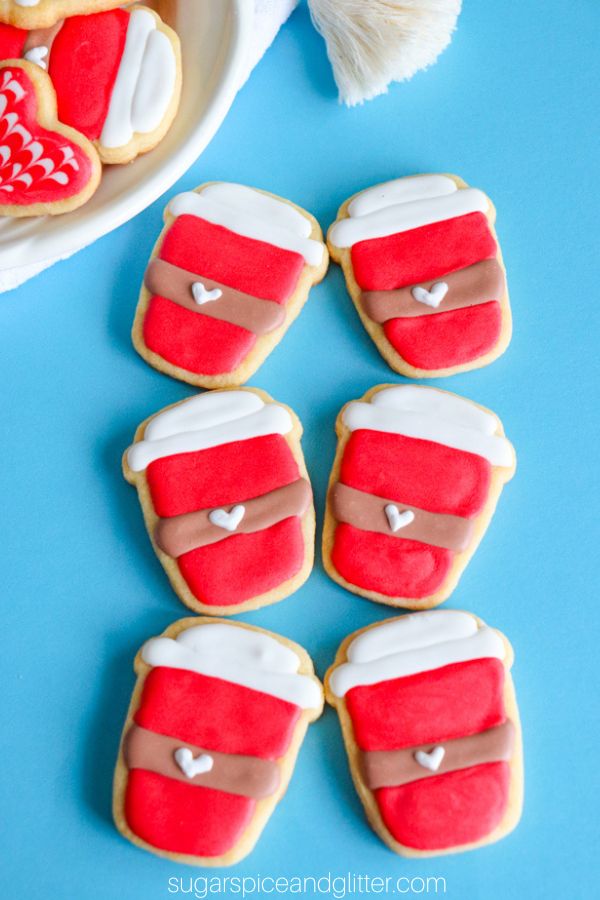six sugar cookies cut in the shape of to-go coffee cups and decorated to look like Starbucks' red holiday cups, set on a blue background