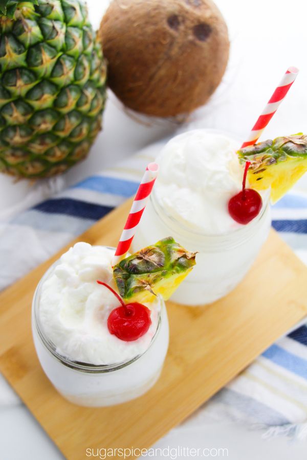overhead image of two mason jars filled with whipped pina colada cocktails topped with pineapple whipped cream and garnished with maraschino cherries and pineapple wedges on a wooden tray with a pineapple and coconut in the background