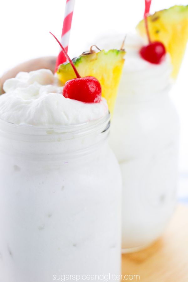 close-up of two mason jars filled with whipped pina colada cocktails topped with pineapple whipped cream and garnished with maraschino cherries and pineapple wedges on a wooden tray