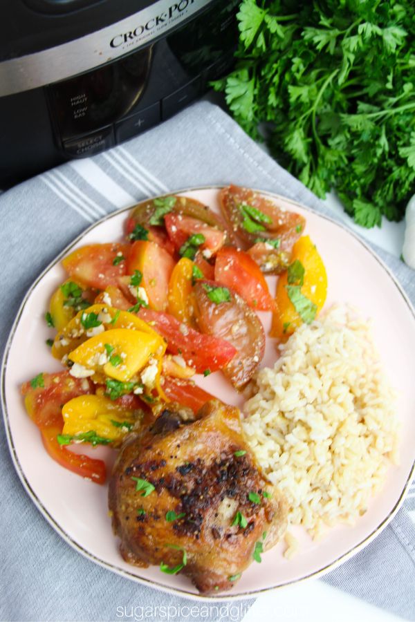 overhead image of a pink plate with brown sugar garlic chicken, rice and a tomato salad in front of a crockpot