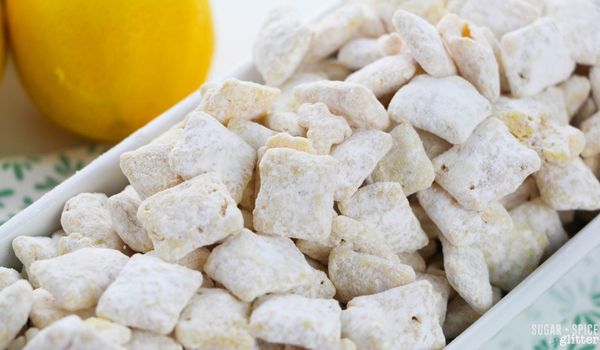 a white bowl filled with lemon muddy buddies on top of a lemon printed napkin with bright yellow lemons in the background