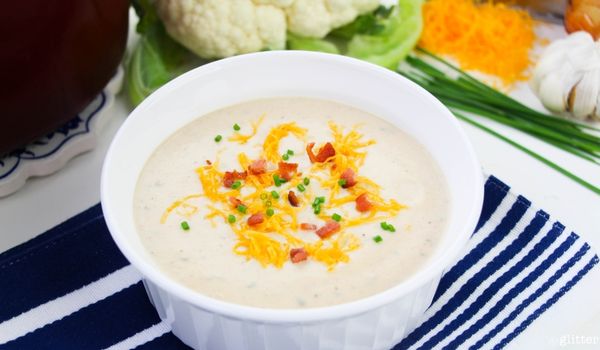 a white bowl full of loaded cauliflower soup garnished with shredded cheddar cheese, bacon bits and chopped chives set on a blue striped placemat, with a head of cauliflower in the background