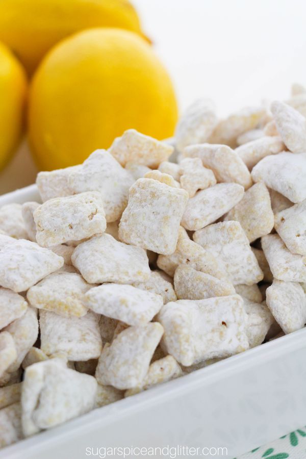 close-up picture of a white bowl filled with lemon muddy buddies with bright yellow lemons in the background