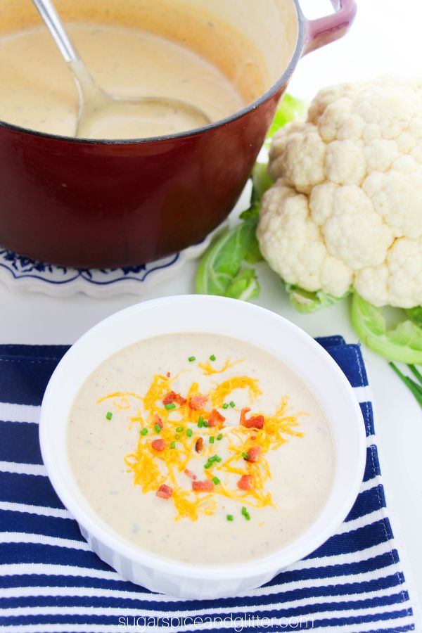 a white bowl full of loaded cauliflower soup garnished with shredded cheddar cheese, bacon bits and chopped chives set on a blue striped placemat, with a head of cauliflower in the background