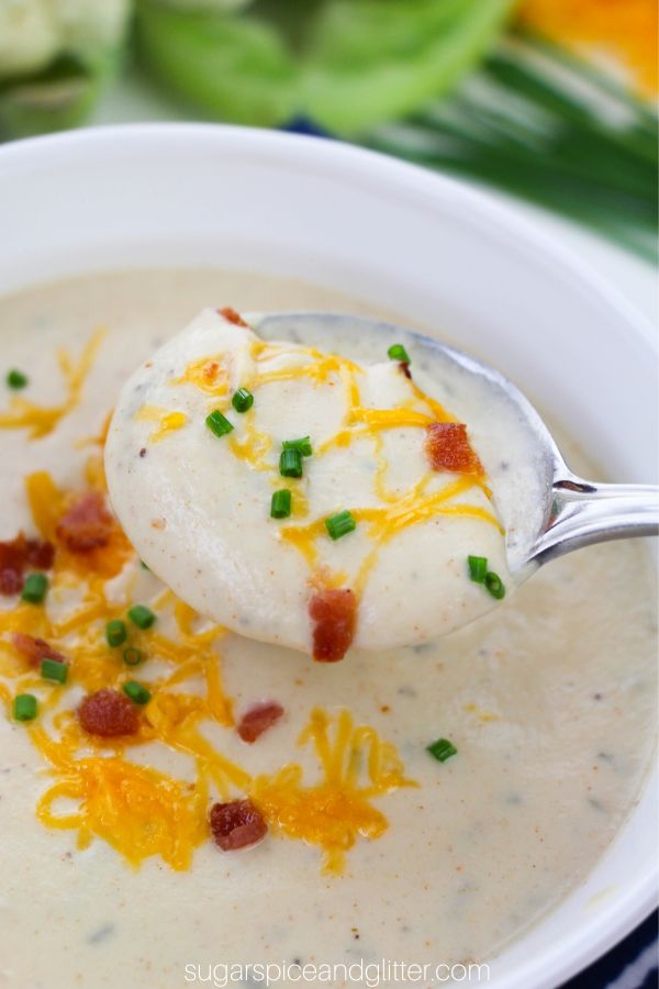 a spoonful of loaded cauliflower soup overtop of a white bowl full of more soup garnished with shredded cheddar cheese, bacon bits and chopped chives