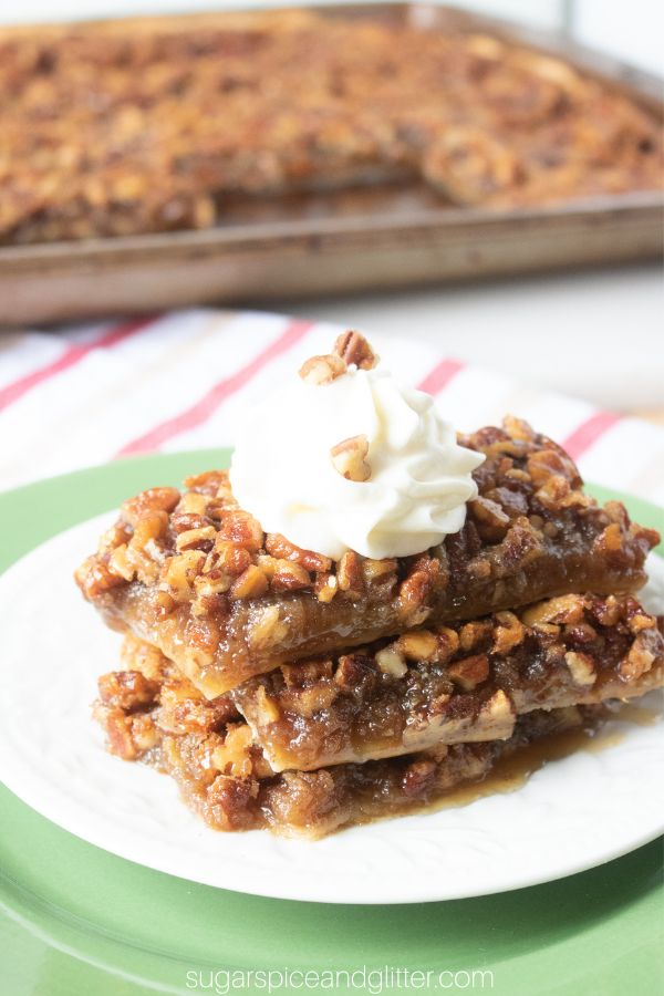 a stack of pecan pie square slices on a white plate, topped with a dollop of whipped cream with a full sheet pan pecan pie in the background