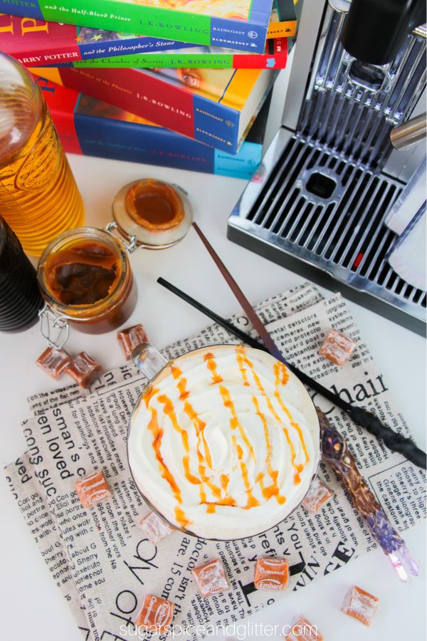 overhead shot of glass mug filled with butterbeer latte on a newsprint napkin with caramel squares and wands scattered around it