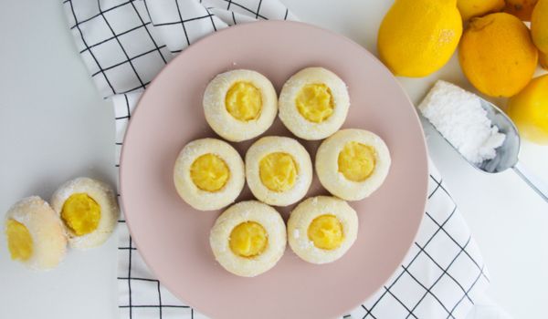 lemon thumbprint cookies on a pink plate with lemons and powdered sugar to the side