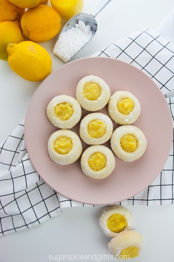 a pink plate holding lemon thumbprint cookies with a bundle of lemons in the corner