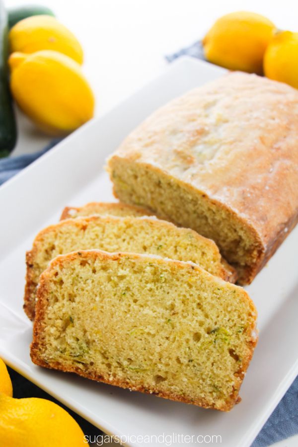 slices of lemon zucchini bread in front of half of a loaf on white platter with lemons and zucchini in the background