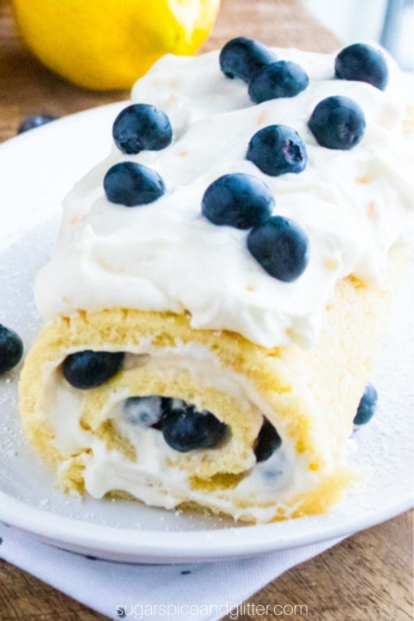 close-up image of a whole lemon jelly cake roll on a white platter slathered with whipped cream and fresh blueberries
