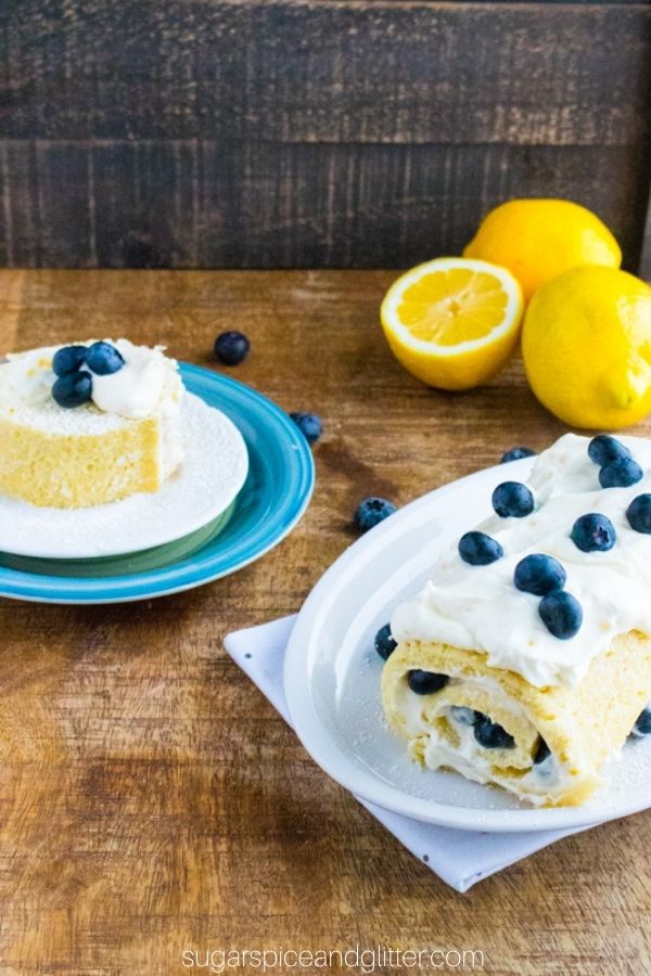 a whole lemon jelly cake roll on a white platter alongside a slice of the cake with lemons and blueberries scattered around