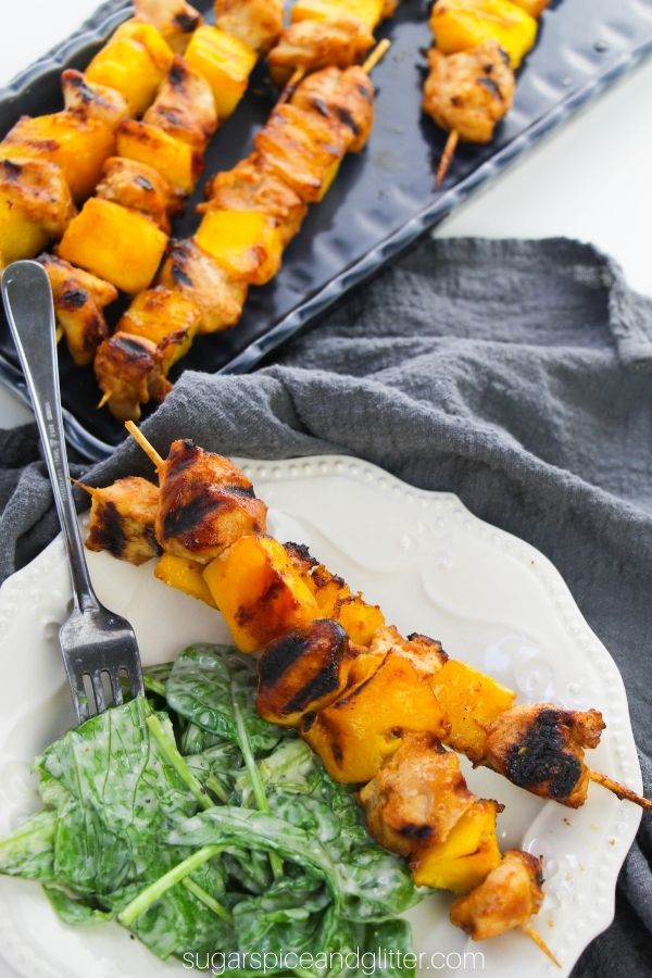 a white plate with two mango chicken kebabs with a side salad, with a blue tray holding more chicken kebabs in the background