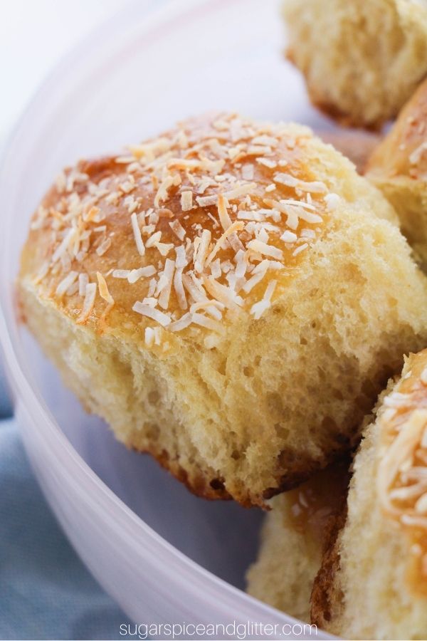 golden Hawaiian dinner rolls garnished with toasted coconut in a pink bowl set on a denim napkin