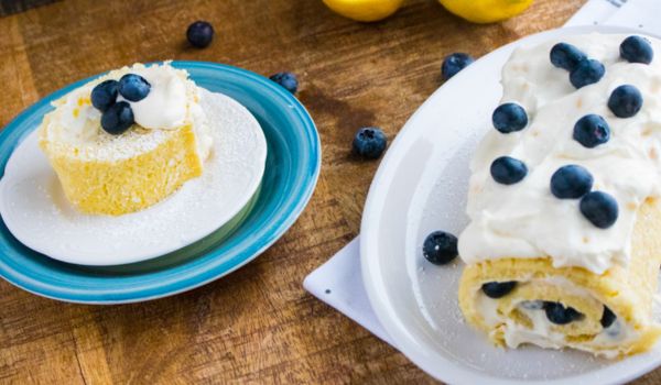 a whole lemon jelly cake roll on a white platter alongside a slice of the cake with lemons and blueberries scattered around