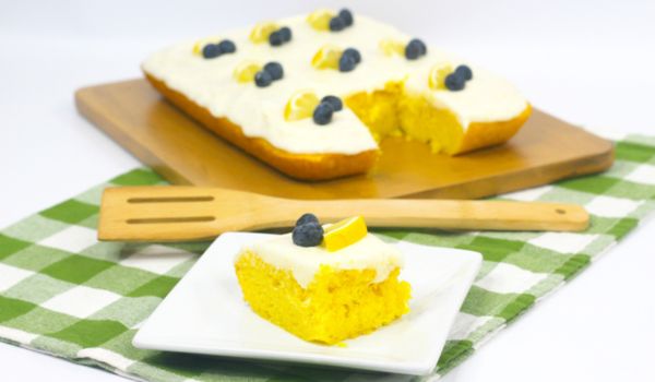 a square of lemon sheet cake on a white plate, set in front of the whole cake on a green plaid napkin