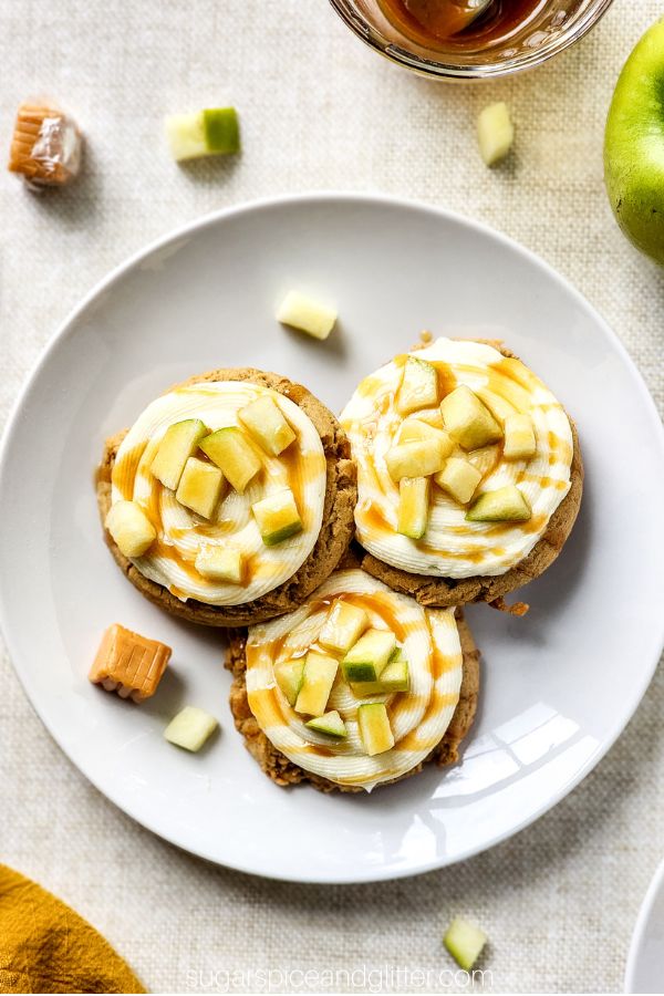 overhead image of caramel apple cookies on a white plate on a linen tablecloth; brown cookies with cream cheese frosting swirled on and Granny Smith apple pieces sprinkled onto the frosting, drizzled all over with caramel