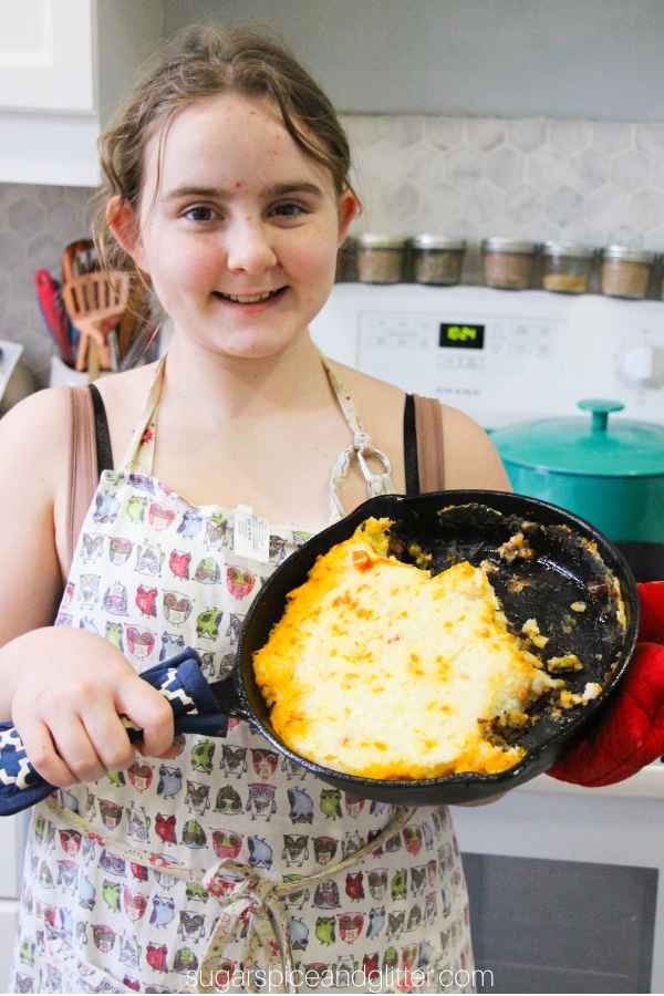 child with hair tied back and wearing an apron, proudly showing off a cottage pie made in a cast iron skillet