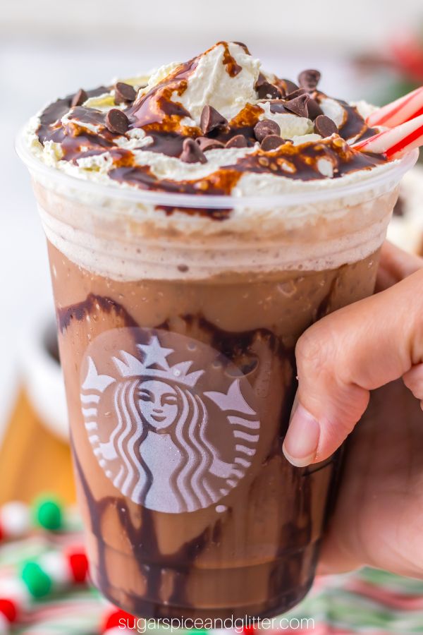 close-up image of a hand holding a clear Starbucks cup holding a peppermint mocha frappuccino topped with whipped cream, chocolate and a candy cane