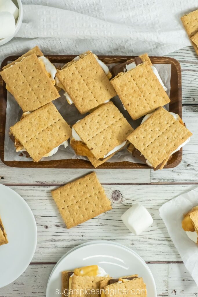 overhead image of a wooden platter of smores, a white plate with a stack of s'mores and s'mores ingredients scattered around