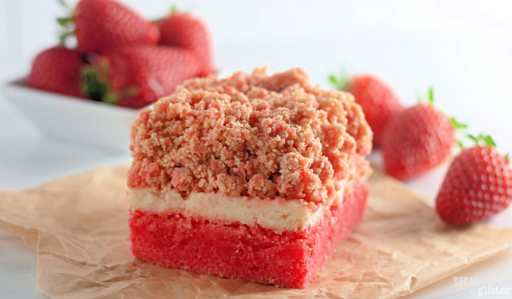 a strawberry crunch cake bar on a brown parchment square with fresh strawberries scattered around it