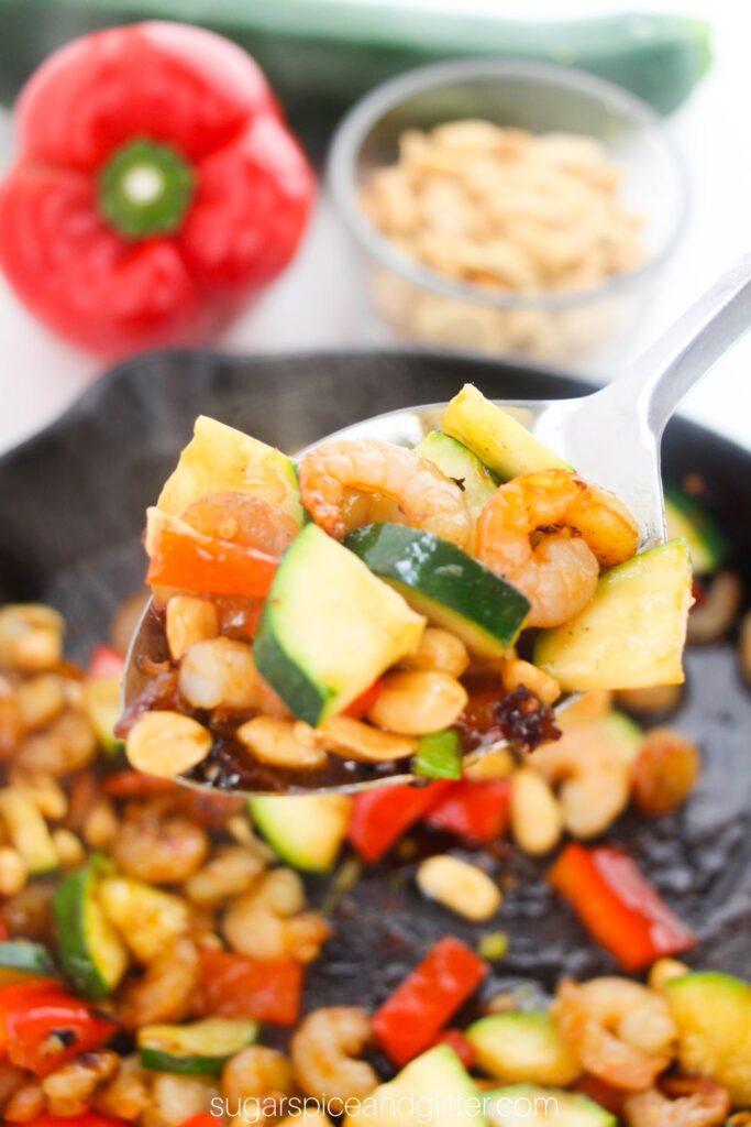 a silver serving spoon holds a scoop of Kung Pao shrimp over a cast iron skillet