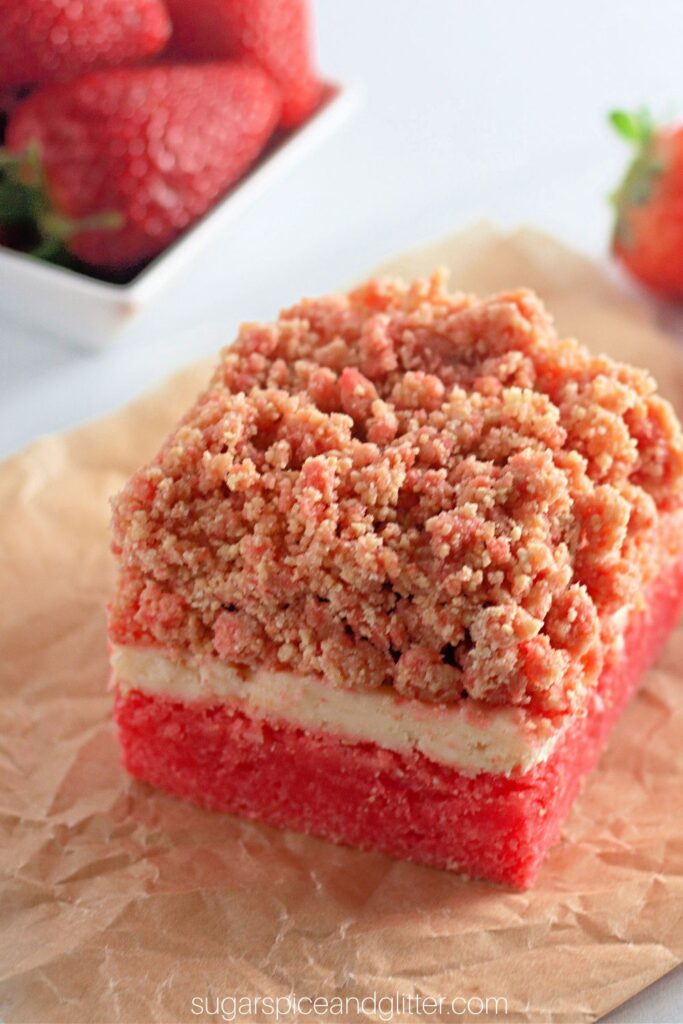 a strawberry crunch cake bar on a brown parchment square with fresh strawberries scattered around it